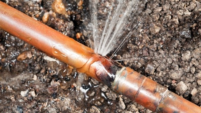 Water dripping from a corroded copper pipe joint beneath lifted floorboards